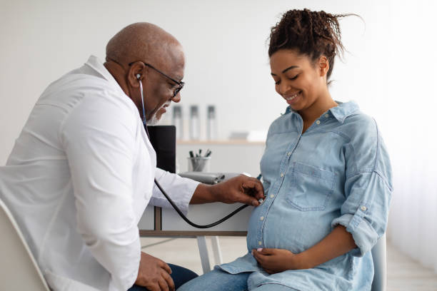 Gynecology consultation showing a smiling Black pregnant woman at a maternity clinic, with a mature male gynecologist examining her belly using a stethoscope during a medical check-up, symbolizing care, pregnancy monitoring, and discussions on fertility success rates.